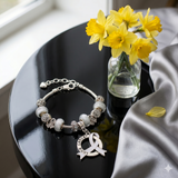Silver bracelet with a heart charm on a black surface with yellow flowers in the background